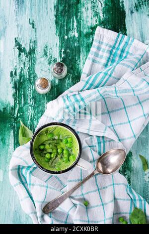 Weißer Vintage-Becher mit Erbsencremesuppe mit Basilikum in Scheiben, serviert mit Löffel, Salz- und Pfefferstreuer auf weißem Küchentuch über grünem und weißem Holz Stockfoto