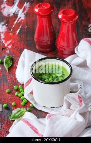 Weißer Vintage-Becher mit Erbsencremesuppe mit Basilikum in Scheiben, serviert mit roten Salz- und Pfefferstreuer auf weißem Küchentuch über rotem und schwarzem Holzrücken Stockfoto