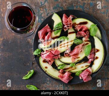 Melone in Scheiben mit Schinken, Basilikumblättern und Balsamico-Sauce, serviert auf schwarzem Teller mit einem Glas Rotwein auf altem, strukturiertem Holzhintergrund. Draufsicht. Wi Stockfoto