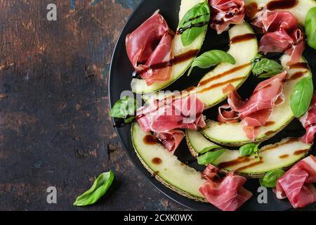 Melone in Scheiben geschnitten mit Schinken und Basilikumblättern, serviert auf schwarzem Keramikplatte über altem Holz texturierten Hintergrund. Draufsicht. Mit Platz für Text Stockfoto