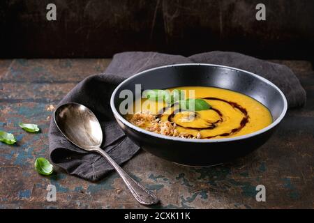 Schwarze Keramikschale aus Kürbis und Süßkartoffel-Sahne-Suppe mit frischem Basilikum, gebratener Zwiebel und Balsamico-Sauce, serviert mit Löffel auf textiler Serviette über o Stockfoto