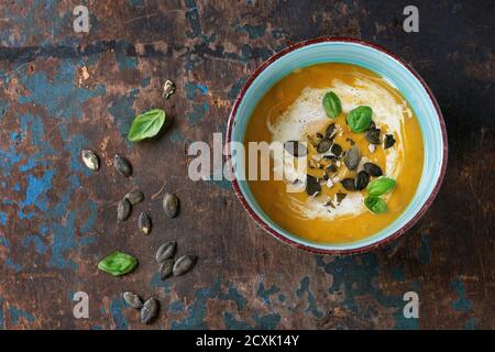 Türkisfarbene Keramikschüssel mit Kürbis und Süßkartoffel-Sahne-Suppe mit frischem Basilikum, Sahne und Samen, serviert auf altem, strukturiertem Holzhintergrund. Draufsicht. Stockfoto