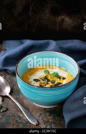 Türkisfarbene Keramikschale aus Kürbis und Süßkartoffel-Sahne-Suppe mit frischem Basilikum, Sahne und Samen, serviert mit Löffel auf textiler Serviette über altem Holz tex Stockfoto