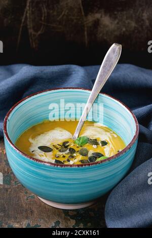 Türkisfarbene Keramikschale aus Kürbis und Süßkartoffel-Sahne-Suppe mit frischem Basilikum, Sahne und Samen, serviert mit Löffel auf textiler Serviette über altem Holz tex Stockfoto