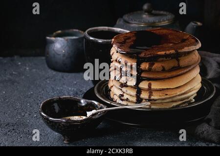 Stapel von hausgemachten amerikanischen Ombre Schokolade Pfannkuchen mit Johannisbrot-Honigsoße serviert auf schwarze Platte mit Krug mit Sahne und Teekanne über schwarze Stein Textur Stockfoto