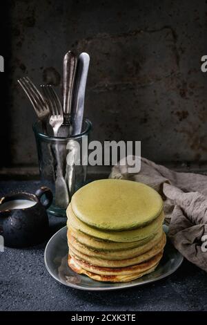 Stapel von hausgemachten american Ombre Green Tea Matcha Pfannkuchen serviert Auf grauem Teller mit Krug Sahne und Besteck in Glas über schwarzem Stein Textur hintergr Stockfoto