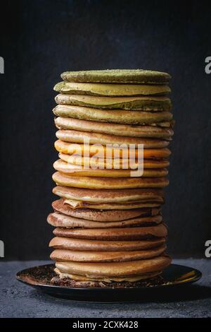 Hohe Stapel von bunten hausgemachte amerikanische Ombre Schokolade, Grüntee Matcha und Kurkuma Pfannkuchen auf Teller über schwarze Steinstruktur Hintergrund. Stockfoto