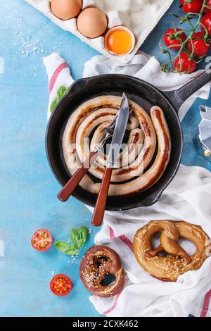 Zutaten zum Frühstück mit Bratwurst in gusseiserner Pfanne, zerbrochenem Ei, Tomaten, Brezeln, Basilikum und Kaffeekanne auf weißem Küchentuch über blauem W Stockfoto