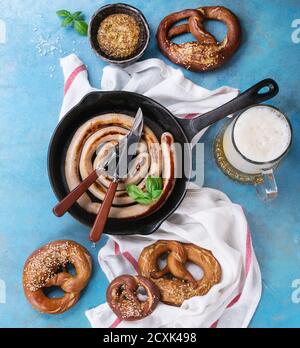 Bratwurst in eiserner Pfanne mit Gabel und Messer, Glas Lagerbier, Senf und Brezeln auf weißem Küchentuch auf blauem Holzhintergrund. Oben Stockfoto