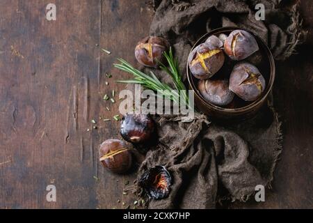 Gebratene Kastanien in der Asche mit Rosmarin in Blechdose auf Sackleinen über alten dunklen hölzernen Hintergrund. Draufsicht mit Platz für Text. Stockfoto