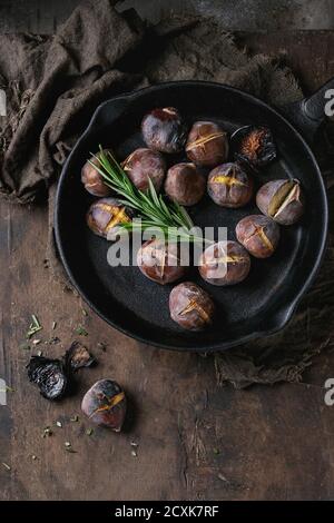 Gebratene Kastanien in der Asche mit Rosmarin in gusseisernen Pfanne auf Sackleinen über alten dunklen hölzernen Hintergrund. Draufsicht mit Platz für Text. Stockfoto