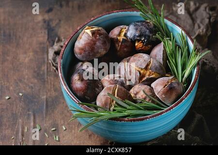 Geröstete Kastanien in der Asche mit Rosmarin in türkisfarbener Keramikschale auf Sacktuch über altem dunklen Holzhintergrund. Nahaufnahme Stockfoto