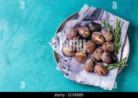 Geröstete Kastanien in der Asche mit Rosmarin in Metallplatte auf Papier auf hellem türkisfarbenem Holzhintergrund. Draufsicht mit Platz für Text. Stockfoto