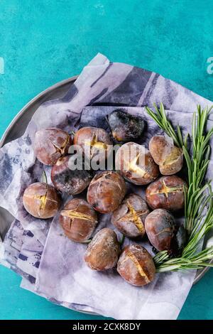 Geröstete Kastanien in der Asche mit Rosmarin in Metallplatte auf Papier auf hellem türkisfarbenem Holzhintergrund. Draufsicht mit Platz für Text. Stockfoto