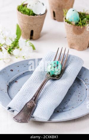 Tischdekor farbenfrohe Osterquaileier mit Frühlingskirschblüten, Moos in Gartentöpfen, leere Teller, Vintage-Gabel über weißer Tischdecke. Stockfoto