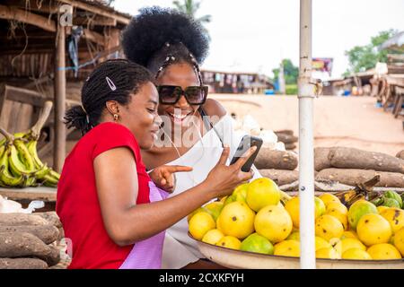 Zwei junge Frauen in einem lokalen Markt, ein Händler und ihr Kunde, die Inhalte auf einem Telefon zusammen ansehen Stockfoto