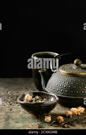 Schwarze Eisen Teekanne mit Keramik Tasse heißen Tee und Vielfalt von schwarzen, grünen und pflanzlichen trockenen Teeblättern und Rosenblüten in Schüssel über alten dunklen Holz backgro Stockfoto