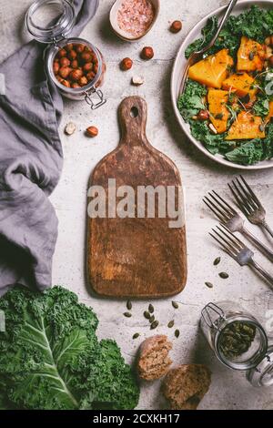 Leere Holzschneidebrett umgeben saisonalen Herbst Produkte.gerösteten Kürbissalat, Grünkohl, Brot, Nüsse und Samen. Draufsicht, Food Frame Hintergrund. Stockfoto