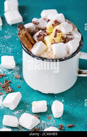 Heiße Schokolade mit hausgemachtem Marshmallow, Schokoladenstückchen, Zimtsticks und Sirup in weißem Vintage Emaille Becher über helltürkisfarbenem Holzhinterrohn Stockfoto