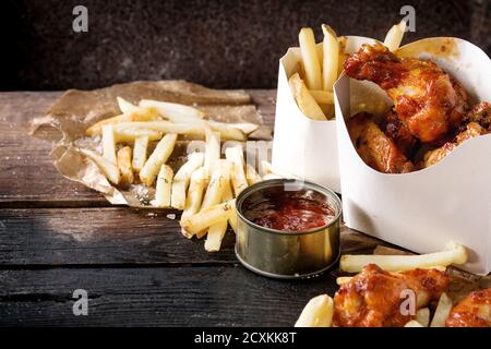 Fast-Food gebraten Huhn Beine, Flügel und Pommes frites Kartoffeln in Lunch-Boxen mit Salz und Ketchup serviert auf Backpapier über alte dunkle wo Soße Stockfoto