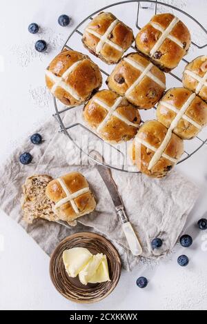 Heiße Kreuzbrötchen auf Backblech serviert mit Butter, frischen Blaubeeren, Messer und Krug Sahne auf textiler Serviette auf weißem Beton-Hintergrund. Stockfoto