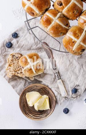 Heiße Kreuzbrötchen auf Backblech serviert mit Butter, frischen Blaubeeren, Messer und Krug Sahne auf textiler Serviette auf weißem Beton-Hintergrund. Stockfoto