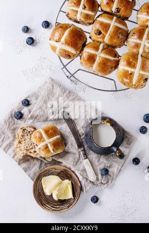 Heiße Kreuzbrötchen auf Backblech serviert mit Butter, frischen Blaubeeren, Messer und Krug Sahne auf textiler Serviette auf weißem Beton-Hintergrund. Stockfoto