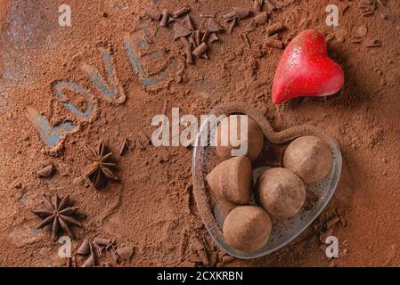 Schokoladentrüffel im Glasherz auf kakaopulvertem Hintergrund mit Valentinskinder, Anissternen und Schriftzug Love. Draufsicht. Geschenk zum Valentinstag Stockfoto