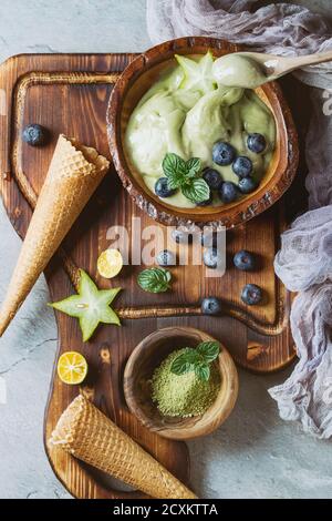 Schale mit grünem Tee Matcha elastische Softeis mit Minze Blätter, Karambolen, Kalk, Heidelbeeren und Waffel Kegel auf hölzernen Portion Board mit Holz spoo Stockfoto