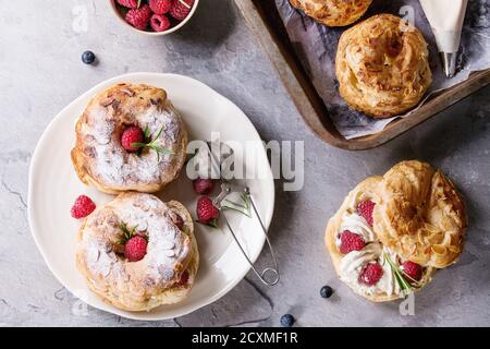 Füllen und leeren hausgemachte Choux Gebäck Kuchen Paris Brest mit Himbeeren, Mandeln, Puderzucker, Rosmarin auf Platte und Ofen Tablett mit Beeren über gra Stockfoto