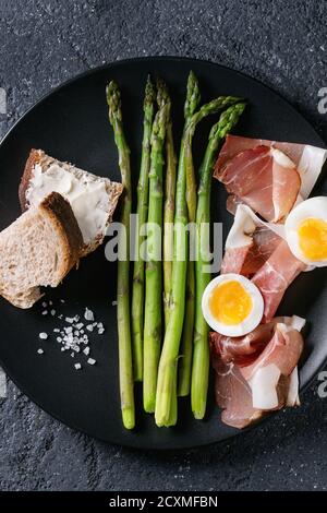 Gekochter grüner Spargel mit halbgekochtem Ei, Brotscheiben und Schinkenspeck, serviert mit Meersalz auf schwarzem Keramikplatte auf dunklem Steintextur-Hintergrund Stockfoto