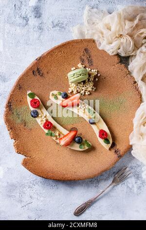 Food Plating Dessert Bio-Banane mit frischen Beeren, Minze, Puffreis und Makronen-Keks serviert mit grünem Tee Matcha-Pulver auf Terrakotta-Platte Stockfoto