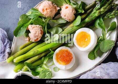 Gekochte grüne Spargel mit gekochtem Ei, Salat, roter Kaviar und Lachs, Pastete auf weißem Fisch Teller serviert mit Gaze Textil über Grau Blau text Stockfoto