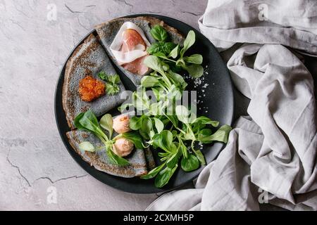 Schwarz Pfannkuchen Crepes mit unterschiedlichsten Salz einfüllen Textile Leinen Serviette über graue Textur staatlich rotem Kaviar, Schinken, Fisch Lachs Pastete, grüner Salat Stockfoto