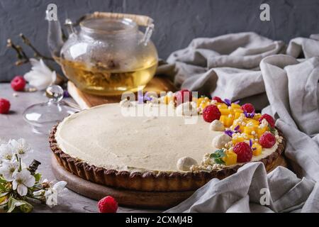 Hausgemachte Schokolade Tarte von Mango, Himbeeren, Minze dekoriert, aufgeblasen Reis und essbaren Blumen serviert mit Glas Teekanne und Textile Leinen über graue tex Stockfoto