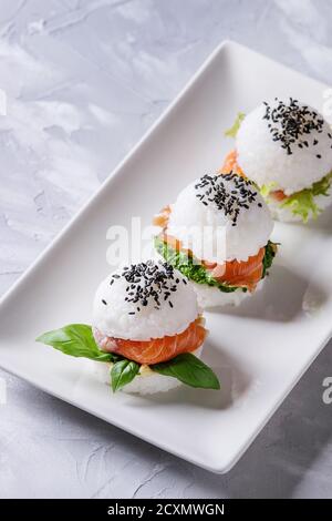 Mini-Reis-Sushi-Burger mit geräuchertem Lachs, grünem Salat und Saucen, schwarzem Sesam serviert auf weißem quadratischem Teller auf grauem Betonboden. Modern HE Stockfoto