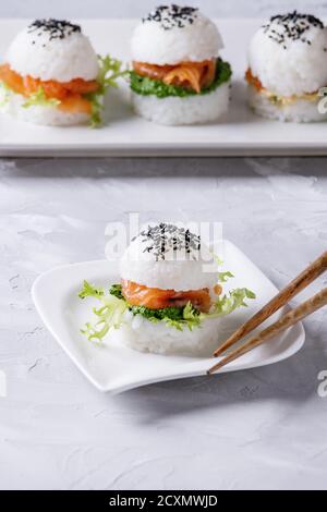 Mini Reis sushi Burger mit geräuchertem Lachs, grüner Salat und Saucen, schwarzer Sesam auf weiße quadratische Platte mit hölzernen Eßstäbchen über Graues konkret serviert. Stockfoto