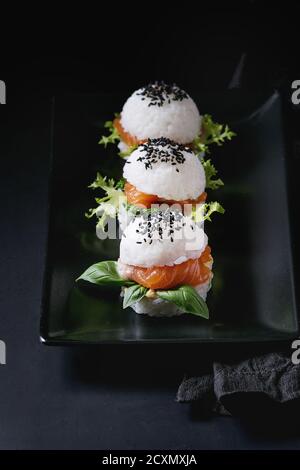 Mini Reis sushi Burger mit geräuchertem Lachs, grüner Salat und Saucen, schwarzer Sesam auf Schwarze quadratische Platte mit textilen Serviette serviert über schwarzen backgroun Stockfoto