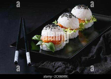 Mini Reis sushi Burger mit geräuchertem Lachs, grüner Salat und Saucen, schwarzer Sesam auf Schwarze quadratische Platte mit Stäbchen und textile Serviette serviert über Stockfoto
