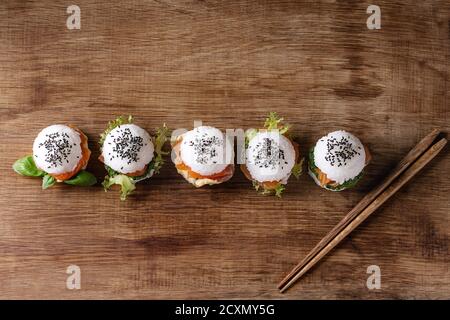Mini Reis sushi Burger mit geräuchertem Lachs, grüner Salat und Saucen, schwarzer Sesam mit Stäbchen aus Holz Über Holz Hintergrund serviert. Moderne gesunde fo Stockfoto