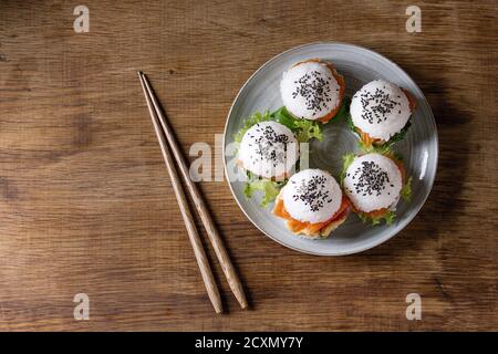 Mini Reis Sushi Burger mit geräuchertem Lachs, grünem Salat und Saucen, schwarzer Sesam serviert auf grauem Teller mit Holzstäbchen auf Holzhintergrund. Mod Stockfoto