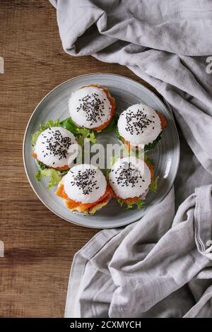 Mini Reis Sushi Burger mit geräuchertem Lachs, grünem Salat und Saucen, schwarzer Sesam serviert auf grauem Teller mit Textilwäsche auf Holzhintergrund. Modern Stockfoto