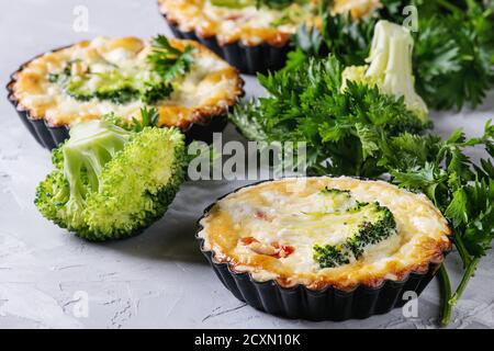 Gebackene hausgemachte Gemüse Brokkoli Quiche Pie in Mini-Metall-Formen serviert mit frischem Grün auf grauem Beton Hintergrund. Nahaufnahme. Bereit zum Essen Stockfoto