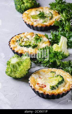 Gebackene hausgemachte Gemüse Brokkoli Quiche Pie in Mini-Metall-Formen serviert mit frischem Grün auf grauem Beton Hintergrund. Nahaufnahme. Bereit zum Essen Stockfoto