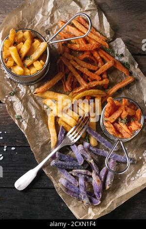 Auswahl an pommes Frites traditionelle Kartoffeln, violette Kartoffeln, Karotten mit Salz, Thymian und Gabel auf Backpapier auf altem Holzhintergrund serviert. Obere V Stockfoto