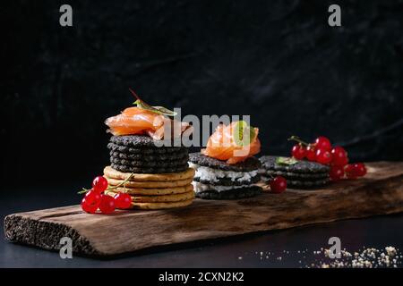 Stapel von schwarzen Holzkohle und traditionellen Cracker mit Räucherlachs, Frischkäse, grüner Salat und rote Johannisbeere Beeren auf Ood dienen Board über schwarz Stockfoto