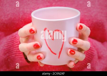 Eine Frau hält eine rosa Tasse mit einem bemalten Herzen. Das Mädchen im roten Pullover mit einem romantischen Becher. Stockfoto