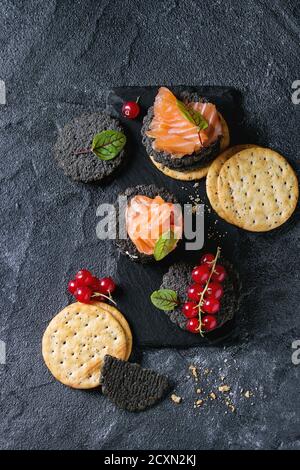 Stapel von schwarzen Vollkorn Holzkohle und traditionellen Cracker mit Räucherlachs, Frischkäse, grüner Salat und rote Johannisbeere Beeren auf Schiefer über Bord Stockfoto