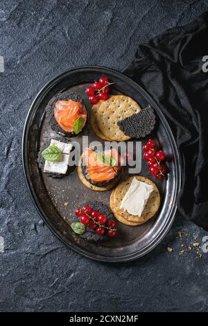 Stapel von schwarzen Holzkohle und traditionellen Cracker mit Räucherlachs, Frischkäse, grüner Salat und rote Johannisbeere Beeren auf Vintage Metalltablett über blac Stockfoto