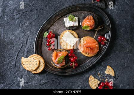 Stapel von schwarzen Holzkohle und traditionellen Cracker mit Räucherlachs, Frischkäse, grüner Salat und rote Johannisbeere Beeren auf Vintage Metalltablett über blac Stockfoto